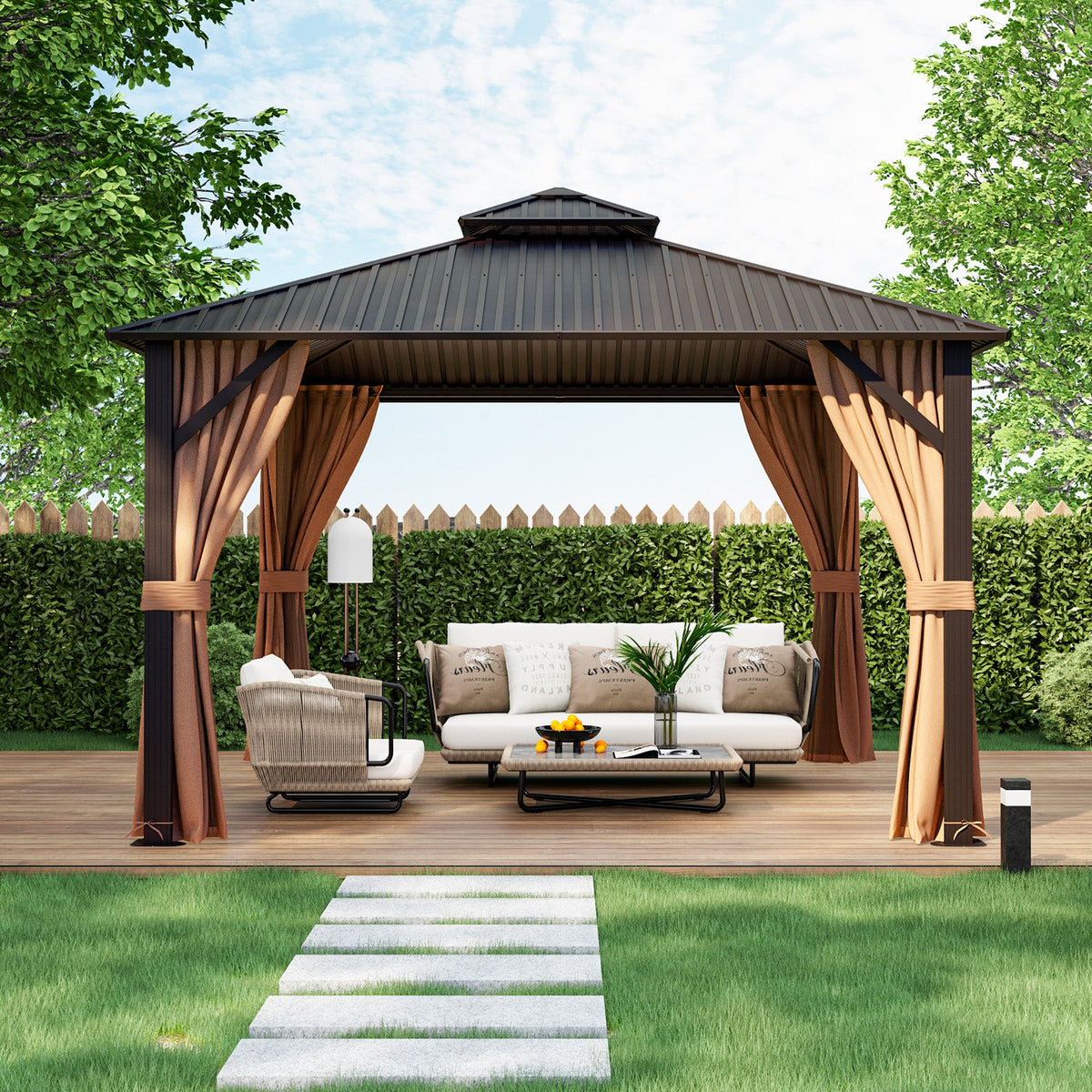 A large outdoor patio gazebo with a dark metal roof and brown curtains frames a white outdoor sofa, a matching armchair, and a coffee table on a wooden deck. The gazebo is set in a backyard with a green hedge, trees, and a grassy lawn with a stone path leading up to it.