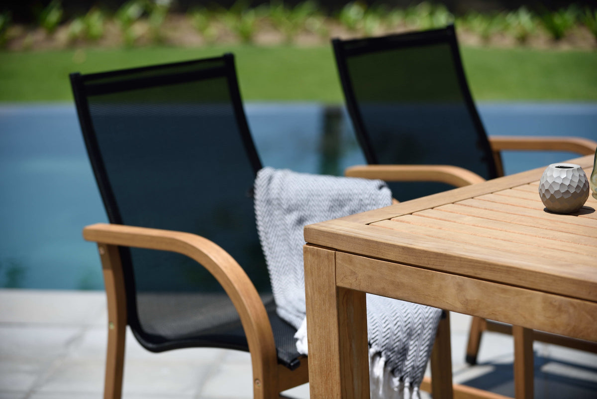 Teak dining table and black mesh armchairs beside a pool.