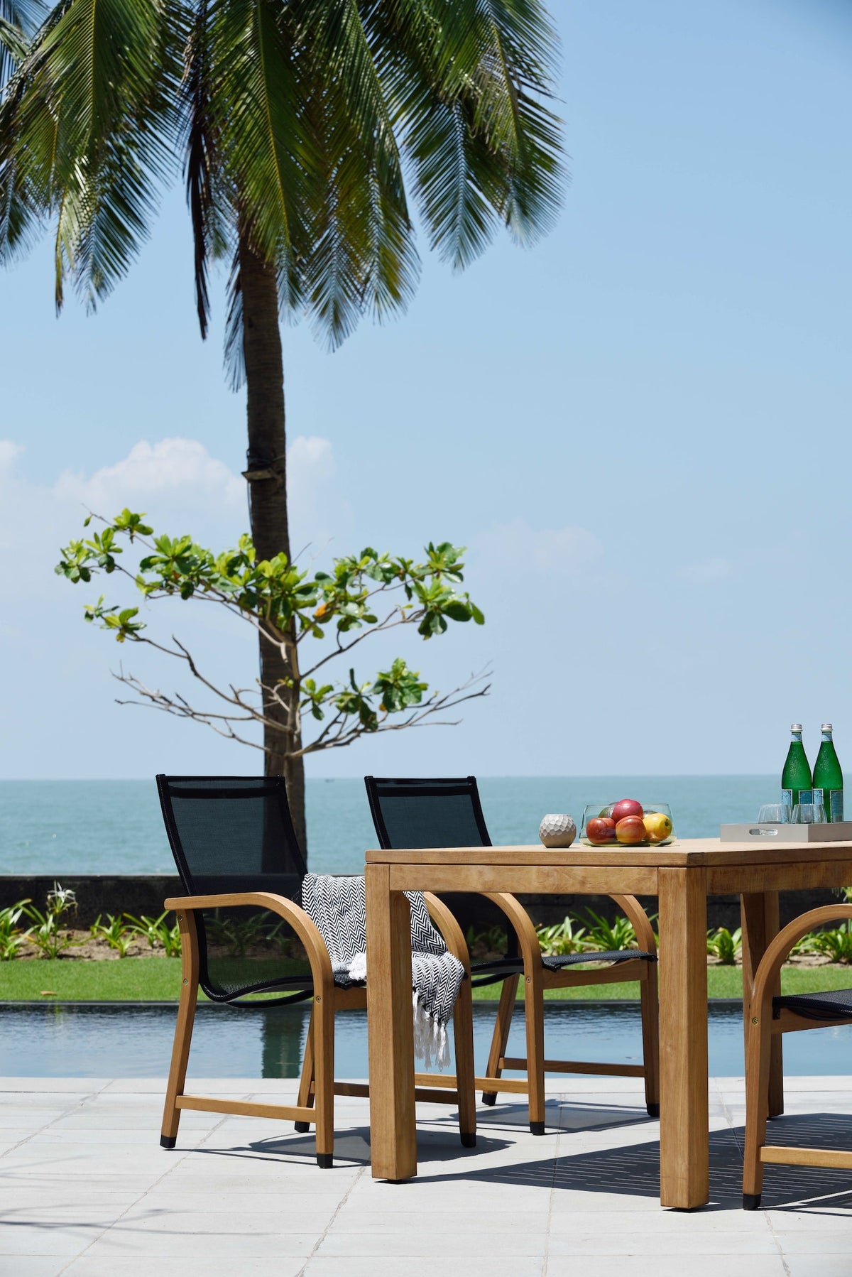 FSC teak dining table with black mesh armchairs beside a pool and ocean view.