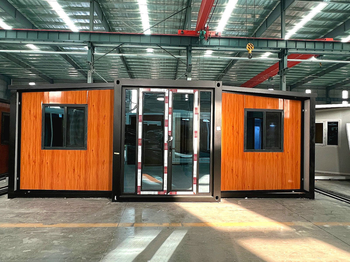 A modular container-style office or cabin with orange wood-look paneling and black framing, shown in a large industrial warehouse with metal roof beams and overhead lighting.