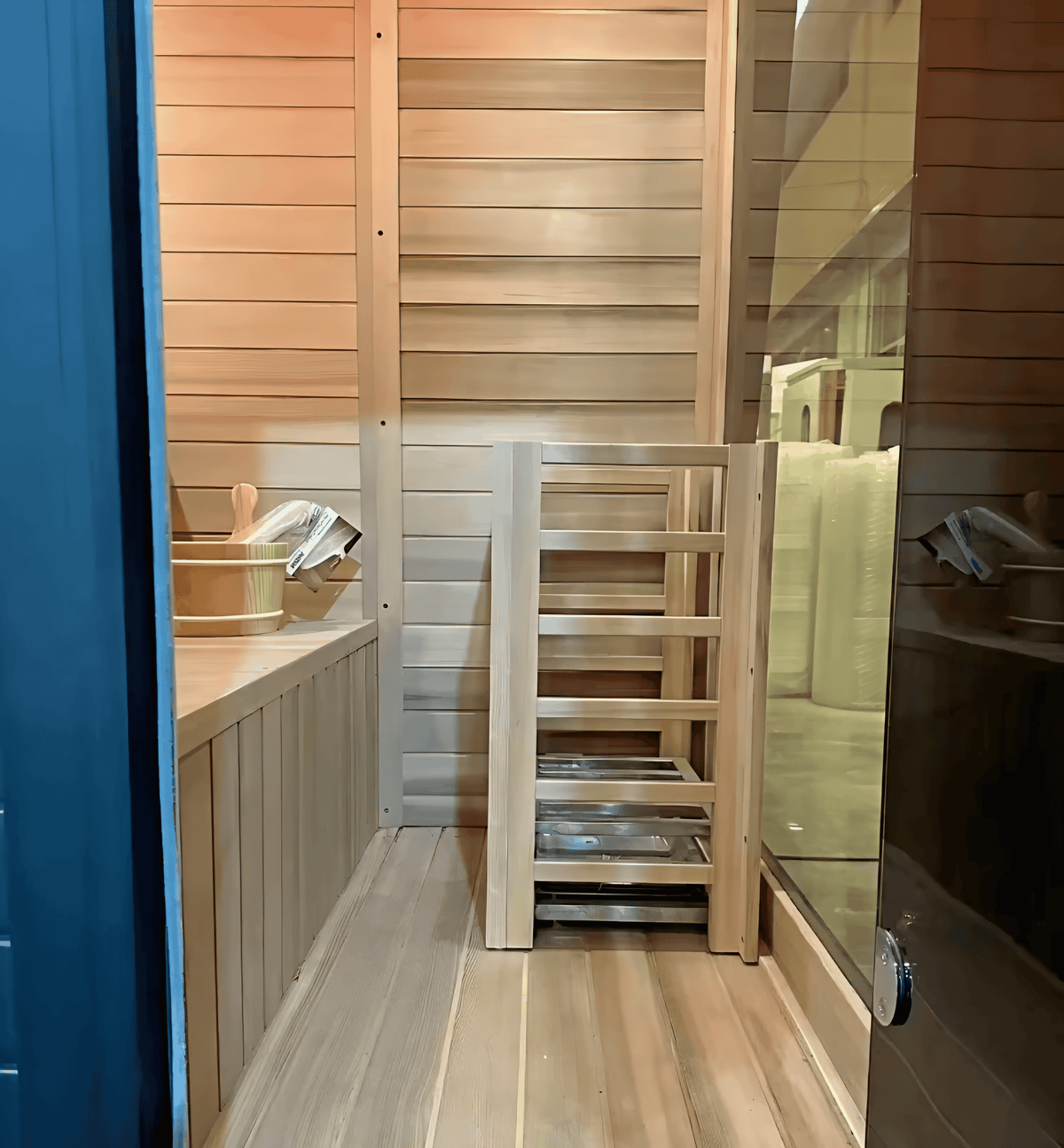 Interior of black hemlock sauna with wooden bench, ladder, and bucket beside the glass door.