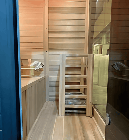 Interior of black hemlock sauna with wooden bench, ladder, and bucket beside the glass door.