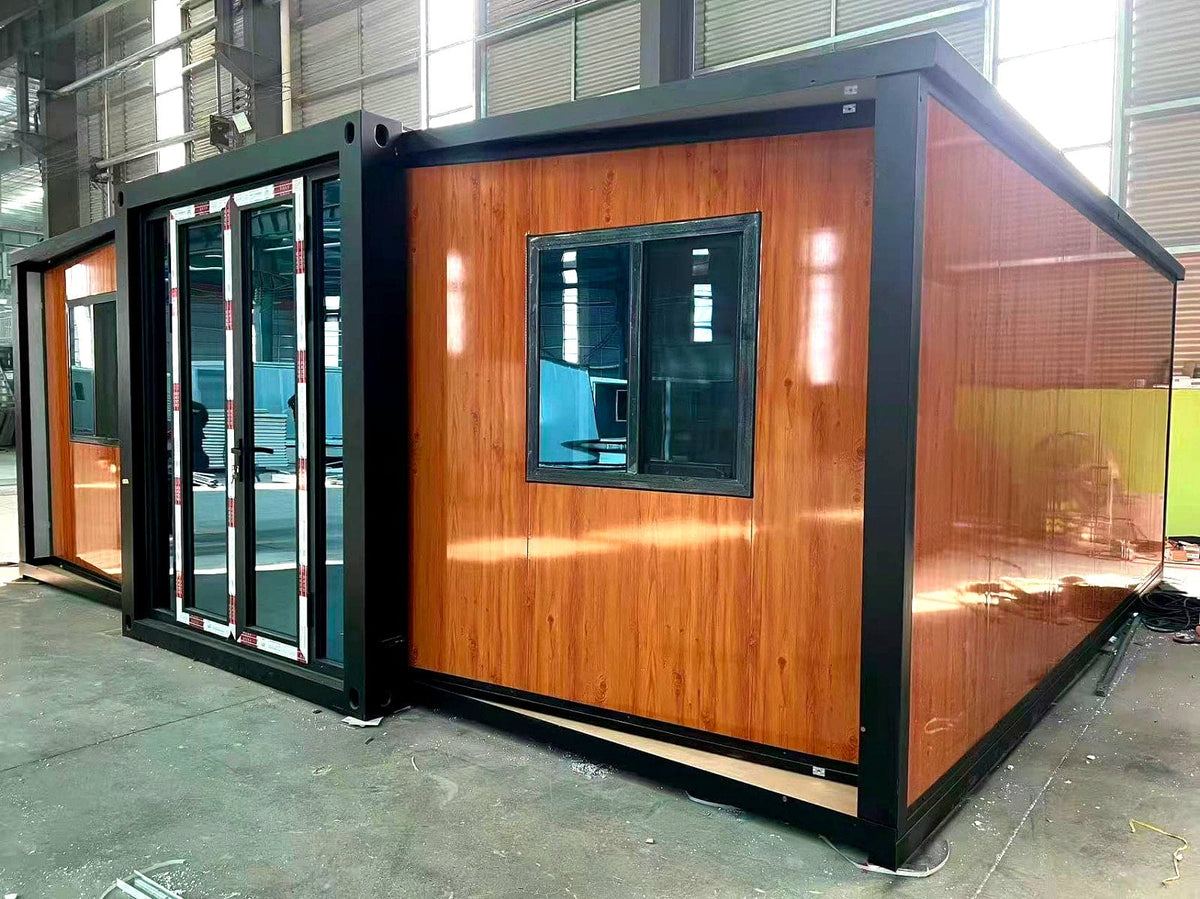 A modular shipping container home with a warm wood-grain exterior paneling and a black metal frame, shown in an industrial warehouse setting.